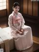 A woman in a pink hanbok sitting on a bed.