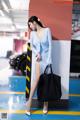 A woman in a blue dress leaning against a wall with a black bag.