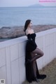 A woman in a black dress leaning against a wall by the ocean.